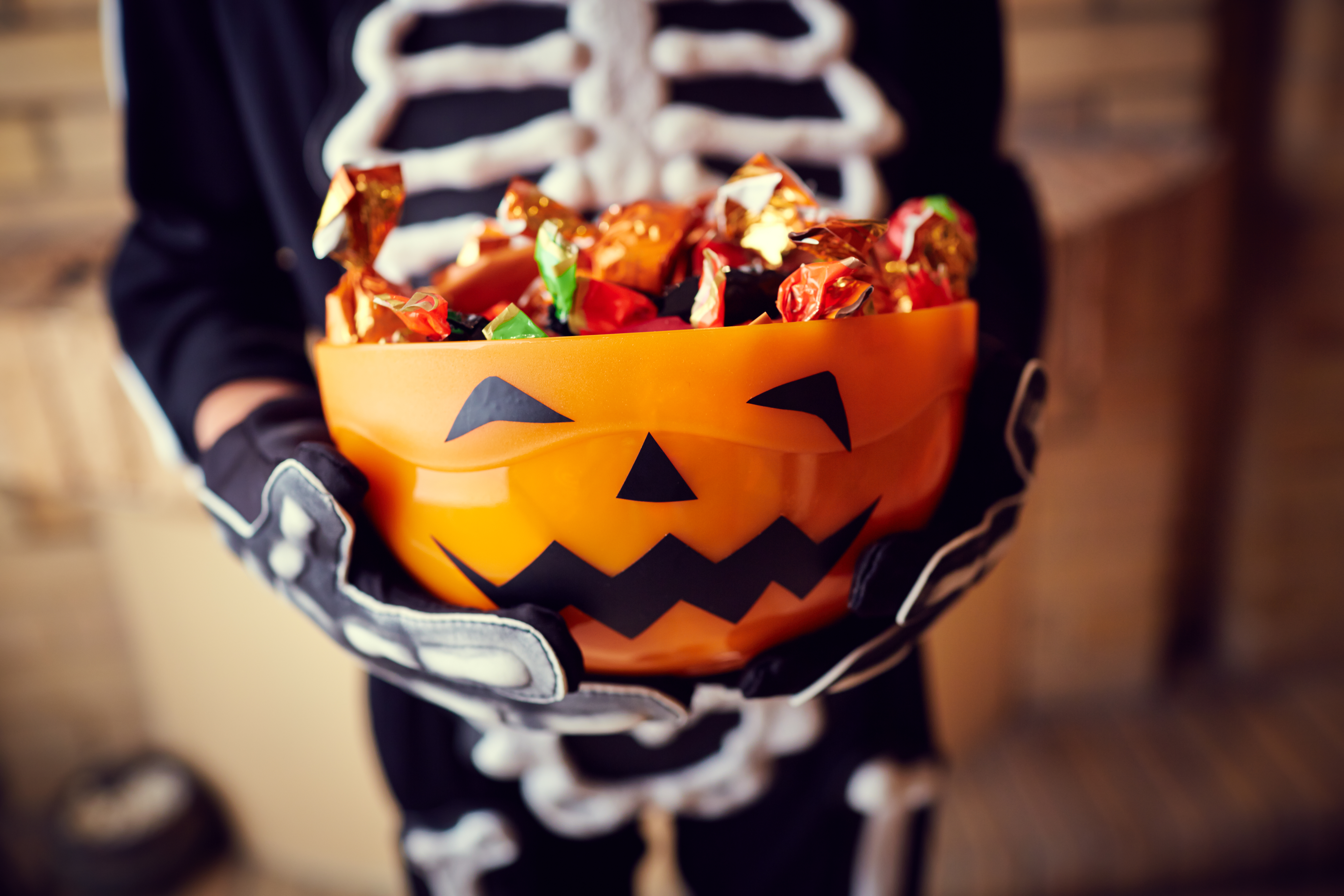 Child in a skeleton costime holding an orange jack-o-lantern bowl filled with candies.
