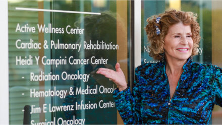 Heidi Y Campini standing in front of the updated Cancer Center entrance, highlighting the door&rsquo;s vinyl lettering reflecting the new name.