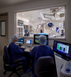 Two staff members at a control console look through a window into one of the two fluoroscopy rooms.